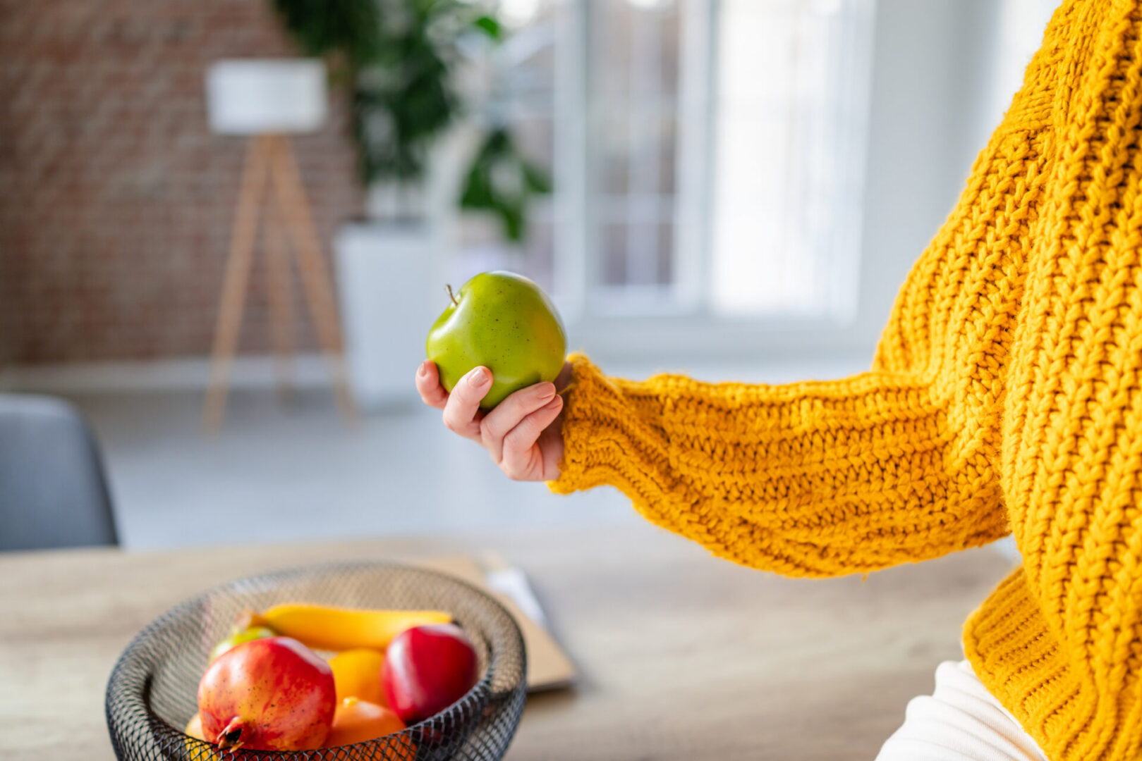 Young woman holds green apple in hand while sitting at table with fruit bowl indoors in bright space during daytime